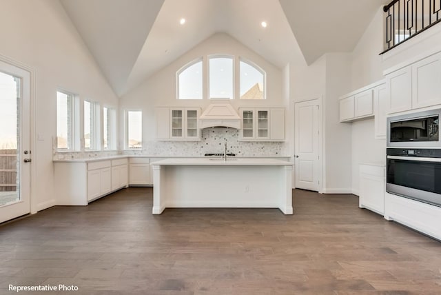 kitchen featuring black microwave, an island with sink, white cabinetry, oven, and custom range hood