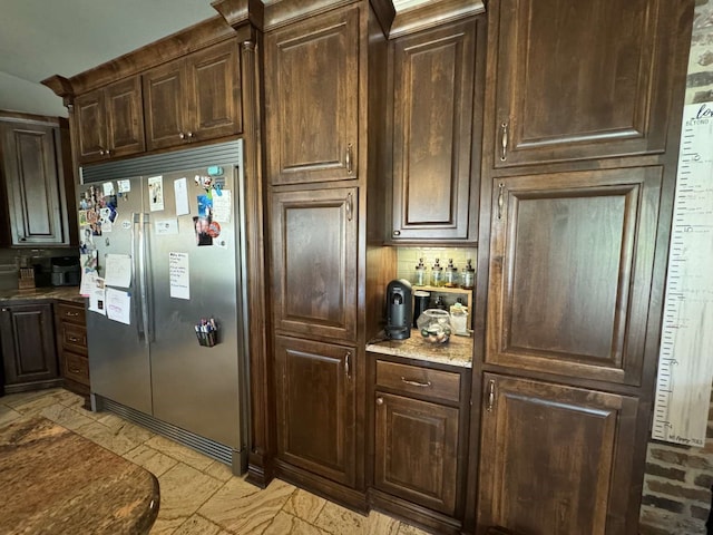 kitchen with light stone countertops, dark brown cabinetry, tasteful backsplash, and built in fridge