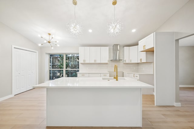 kitchen with wall chimney range hood, white cabinetry, decorative light fixtures, and an island with sink