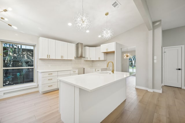 kitchen featuring wall chimney range hood, an island with sink, sink, white cabinets, and light hardwood / wood-style floors