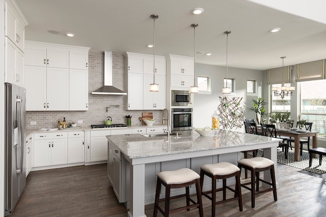 kitchen with wall chimney range hood, white cabinetry, a kitchen island with sink, and appliances with stainless steel finishes