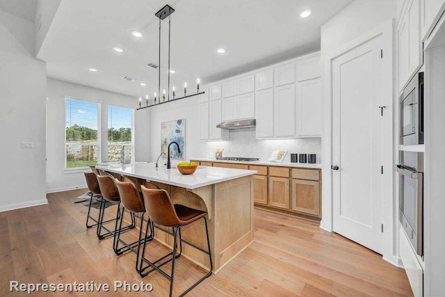 kitchen with stainless steel appliances, a center island with sink, pendant lighting, white cabinets, and light hardwood / wood-style floors