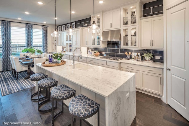 kitchen featuring stainless steel gas cooktop, a center island with sink, light stone counters, and ventilation hood