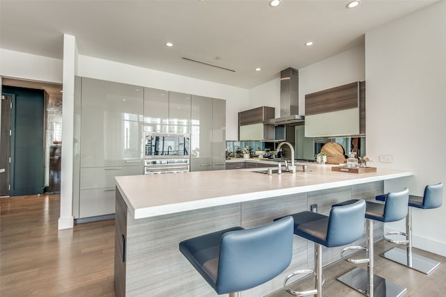 kitchen with wall chimney range hood, oven, kitchen peninsula, light hardwood / wood-style floors, and a breakfast bar area