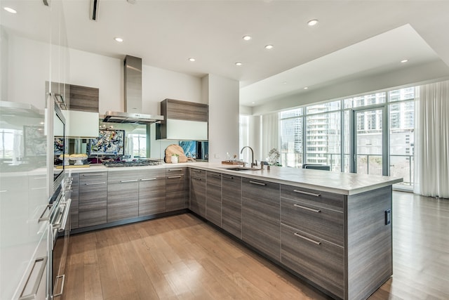 kitchen featuring stainless steel gas cooktop, wall chimney exhaust hood, kitchen peninsula, sink, and light wood-type flooring