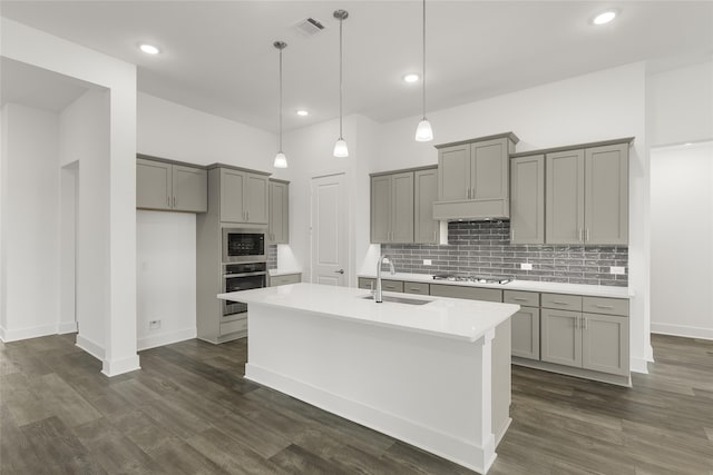 kitchen with sink, an island with sink, hanging light fixtures, stainless steel appliances, and dark wood-type flooring