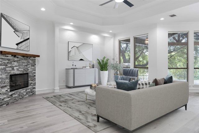 living room featuring ceiling fan, a stone fireplace, light hardwood / wood-style flooring, crown molding, and a tray ceiling