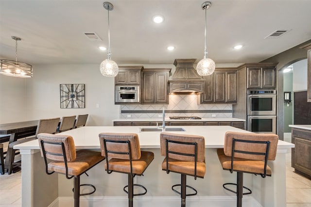 kitchen with dark brown cabinetry, a kitchen breakfast bar, a kitchen island with sink, custom range hood, and appliances with stainless steel finishes