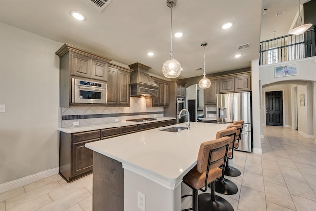 kitchen with a center island with sink, arched walkways, custom range hood, appliances with stainless steel finishes, and light countertops