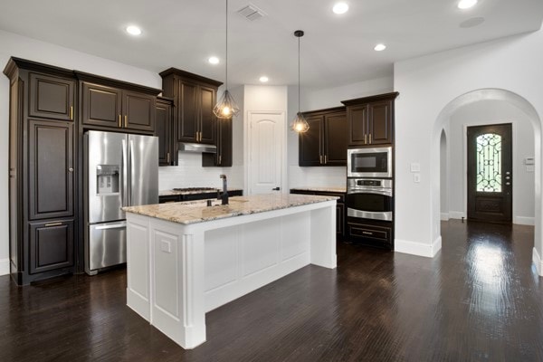 kitchen featuring an island with sink, stainless steel appliances, dark wood-type flooring, pendant lighting, and light stone counters