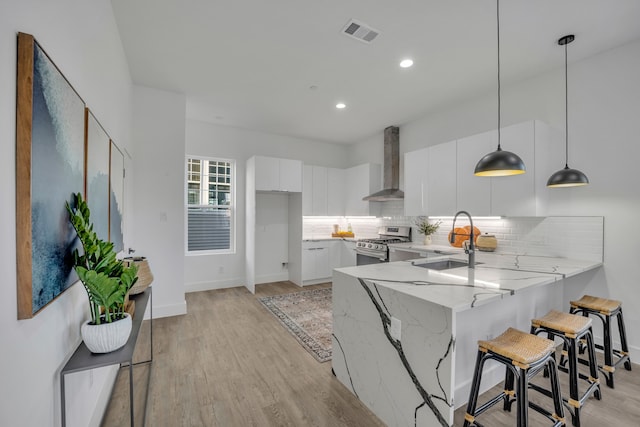 kitchen featuring white cabinets, kitchen peninsula, hanging light fixtures, wall chimney exhaust hood, and stainless steel gas range oven