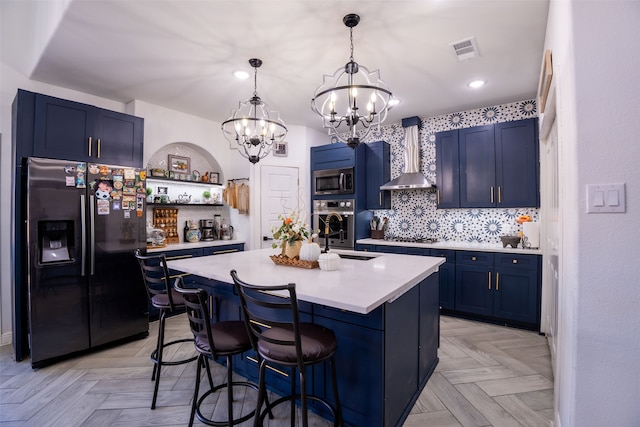 kitchen with tasteful backsplash, wall chimney range hood, appliances with stainless steel finishes, blue cabinetry, and a kitchen island with sink