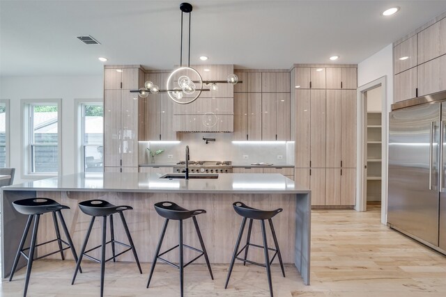 kitchen featuring built in fridge, light hardwood / wood-style flooring, a center island with sink, decorative light fixtures, and light brown cabinetry