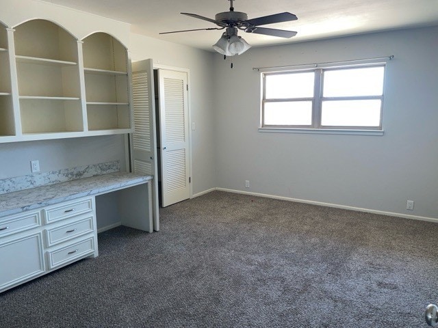 unfurnished office featuring built in desk, dark colored carpet, and ceiling fan