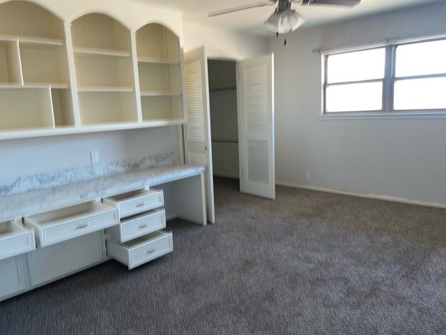 unfurnished office featuring ceiling fan, built in desk, and dark colored carpet