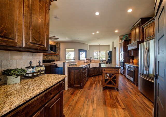 kitchen with stainless steel appliances, tasteful backsplash, light stone counters, dark hardwood / wood-style floors, and an island with sink