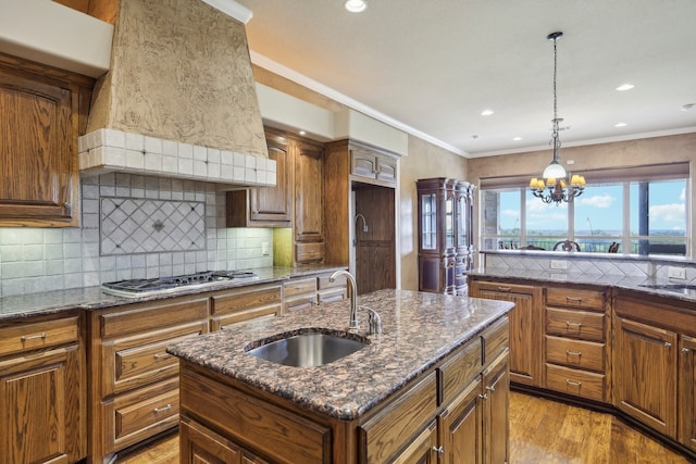 kitchen featuring a center island with sink, dark stone countertops, custom range hood, stainless steel gas cooktop, and sink