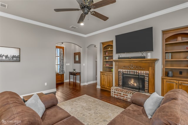 living room with a fireplace, ceiling fan, dark wood-type flooring, crown molding, and built in shelves