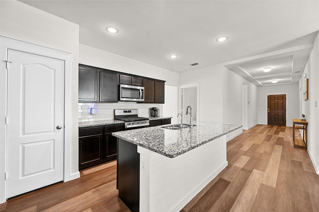 kitchen featuring stainless steel appliances, an island with sink, light stone countertops, sink, and tasteful backsplash