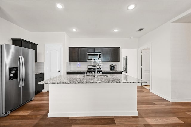 kitchen featuring light stone countertops, a center island with sink, backsplash, appliances with stainless steel finishes, and sink