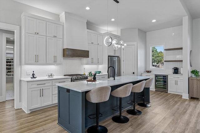 kitchen featuring a center island with sink, beverage cooler, custom exhaust hood, white cabinetry, and light hardwood / wood-style floors