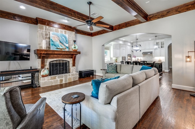 living room featuring ceiling fan with notable chandelier, beam ceiling, a stone fireplace, and dark wood-type flooring