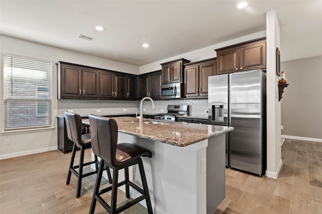 kitchen with a center island with sink, light stone countertops, light wood-type flooring, dark brown cabinetry, and stainless steel appliances