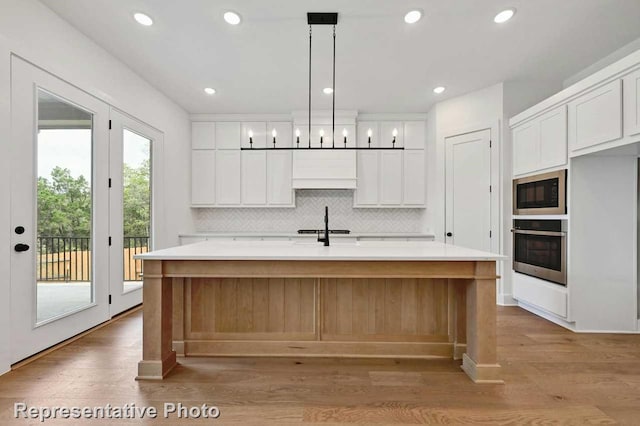 kitchen with a kitchen island with sink, oven, light hardwood / wood-style flooring, hanging light fixtures, and white cabinets