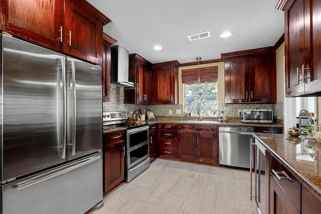 kitchen featuring pendant lighting, dark stone counters, sink, wall chimney exhaust hood, and stainless steel appliances