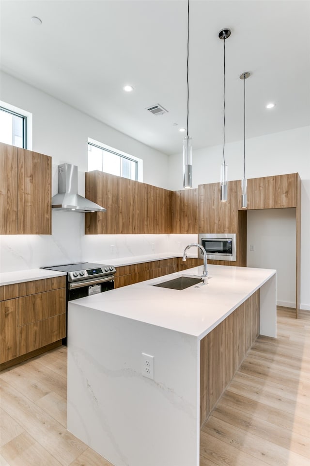 kitchen featuring wall chimney range hood, sink, light wood-type flooring, decorative light fixtures, and a center island with sink