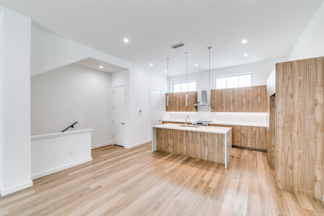 kitchen with light hardwood / wood-style floors, wall chimney exhaust hood, decorative light fixtures, and a kitchen island with sink