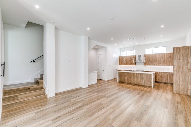 kitchen featuring a large island with sink, wall chimney range hood, hanging light fixtures, sink, and light hardwood / wood-style floors