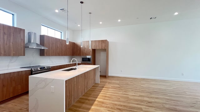 kitchen with a center island with sink, light hardwood / wood-style flooring, stainless steel appliances, wall chimney exhaust hood, and decorative light fixtures