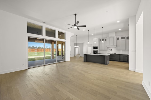 kitchen with light wood-type flooring, ceiling fan with notable chandelier, stainless steel appliances, a kitchen island with sink, and hanging light fixtures