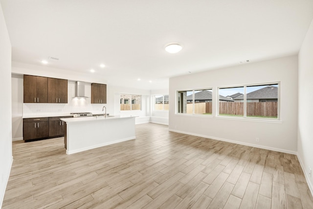 kitchen with wall chimney exhaust hood, light hardwood / wood-style floors, dark brown cabinets, and a kitchen island with sink