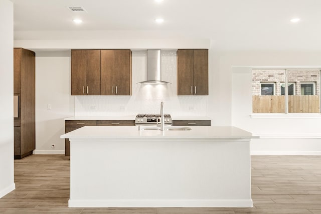 kitchen featuring sink, wall chimney range hood, an island with sink, and light wood-type flooring