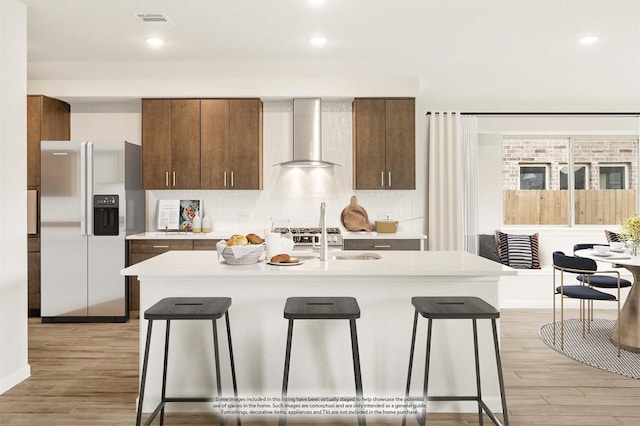 kitchen with wall chimney exhaust hood, stainless steel fridge with ice dispenser, a breakfast bar area, and a kitchen island with sink