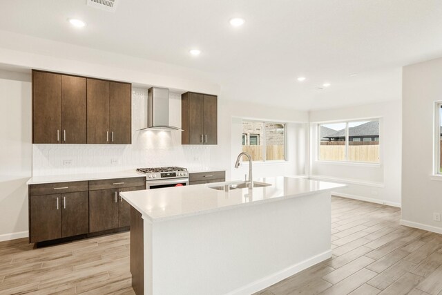 kitchen featuring stainless steel gas range oven, sink, wall chimney exhaust hood, and a center island with sink