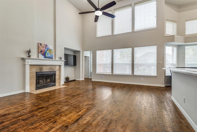 unfurnished living room featuring a tiled fireplace, a towering ceiling, dark wood-type flooring, and ceiling fan