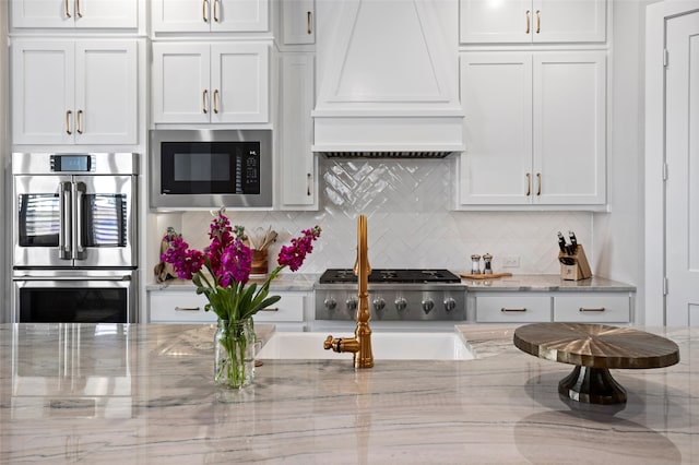 kitchen featuring tasteful backsplash, light stone countertops, appliances with stainless steel finishes, white cabinetry, and premium range hood