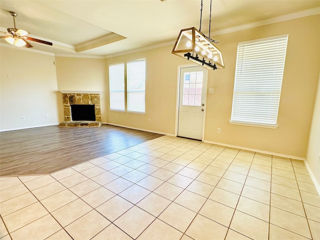 unfurnished living room featuring ceiling fan, a tray ceiling, a stone fireplace, crown molding, and light wood-type flooring