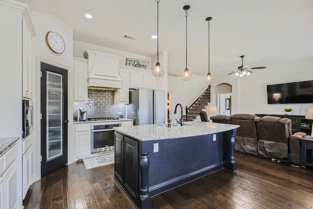 kitchen with light stone counters, a kitchen island with sink, custom range hood, sink, and stainless steel appliances