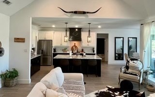 kitchen with a kitchen island with sink, hanging light fixtures, stainless steel fridge, range hood, and white cabinetry