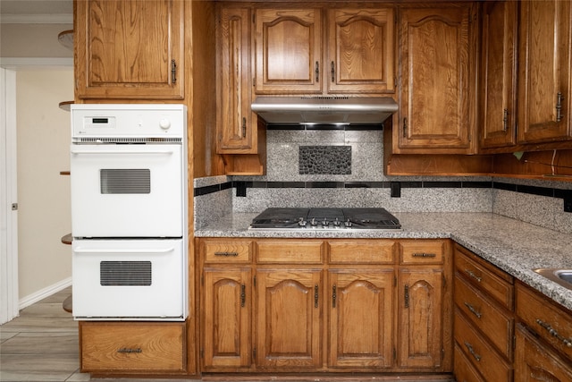 kitchen featuring backsplash, crown molding, stainless steel gas stovetop, light stone counters, and white double oven