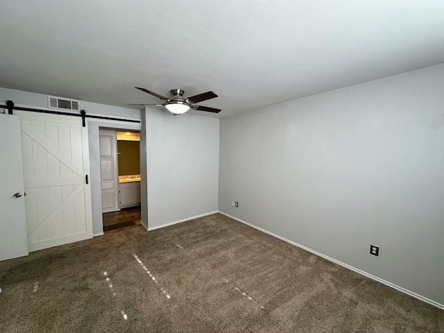 unfurnished bedroom with connected bathroom, ceiling fan, dark colored carpet, and a barn door