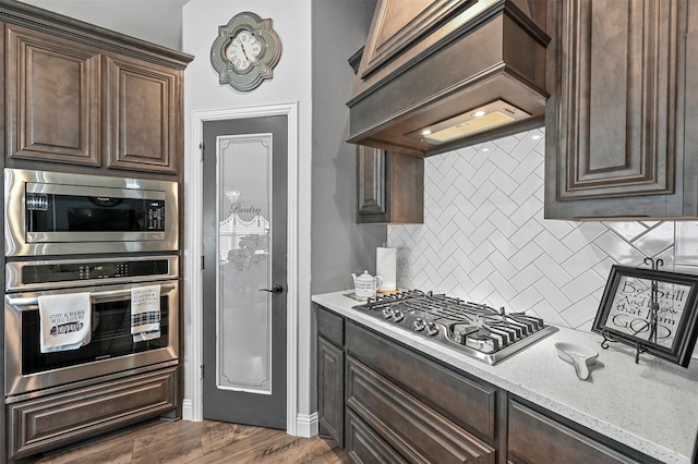 kitchen featuring tasteful backsplash, dark brown cabinetry, custom range hood, light stone counters, and appliances with stainless steel finishes
