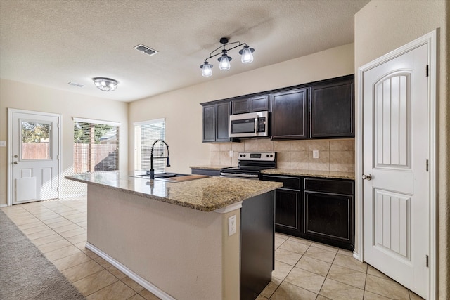 kitchen with appliances with stainless steel finishes, sink, decorative backsplash, light tile patterned floors, and a kitchen island with sink