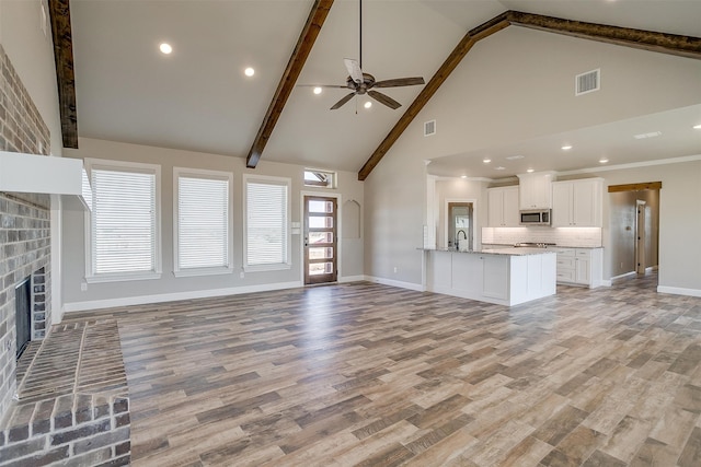 unfurnished living room with beam ceiling, a brick fireplace, light wood-type flooring, high vaulted ceiling, and ceiling fan