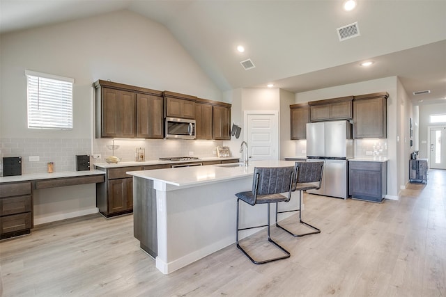 kitchen featuring tasteful backsplash, appliances with stainless steel finishes, light wood-type flooring, and a kitchen island with sink