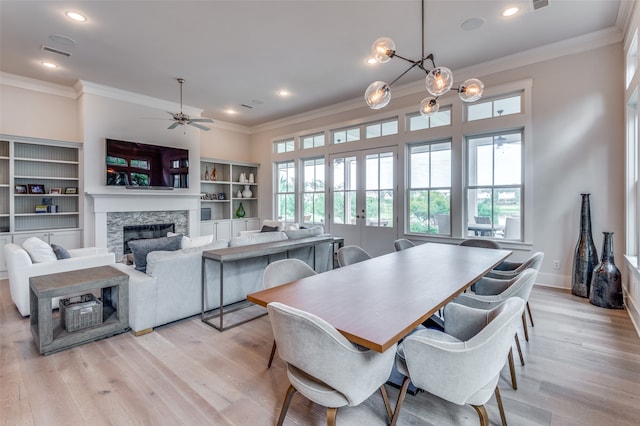 dining space featuring ornamental molding, a stone fireplace, ceiling fan with notable chandelier, and light wood-type flooring